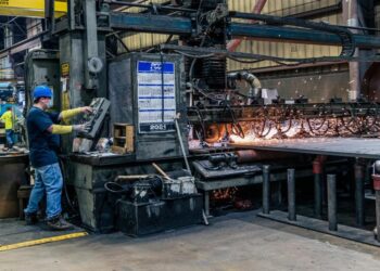 A male engineer wearing a white hard hat and a navy blue industrial uniform operates a touchscreen control panel at a workstation.