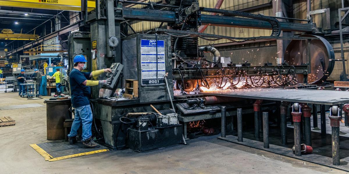 A male engineer wearing a white hard hat and a navy blue industrial uniform operates a touchscreen control panel at a workstation.