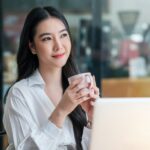 a lady in white holding her cup of coffee in front of her laptop, thinking.