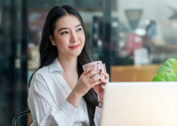 a lady in white holding her cup of coffee in front of her laptop, thinking.