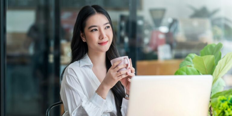 a lady in white holding her cup of coffee in front of her laptop, thinking.